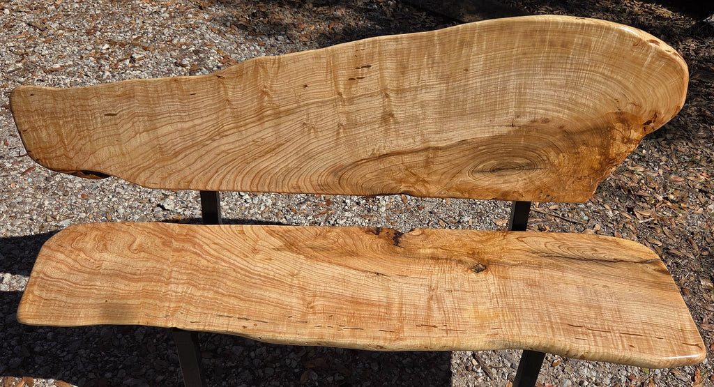 A rustic Ash live-edge wooden bench with a thick natural wood backrest and seat supported by black metal legs, photographed outdoors on gravel.