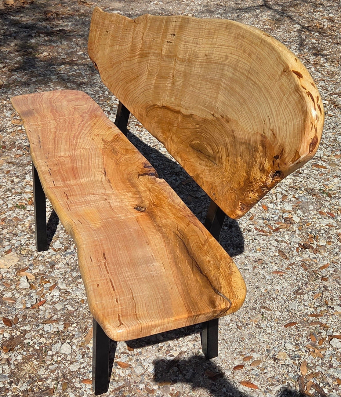 A rustic Ash live-edge wooden bench with a thick natural wood backrest and seat supported by black metal legs, photographed outdoors on gravel.