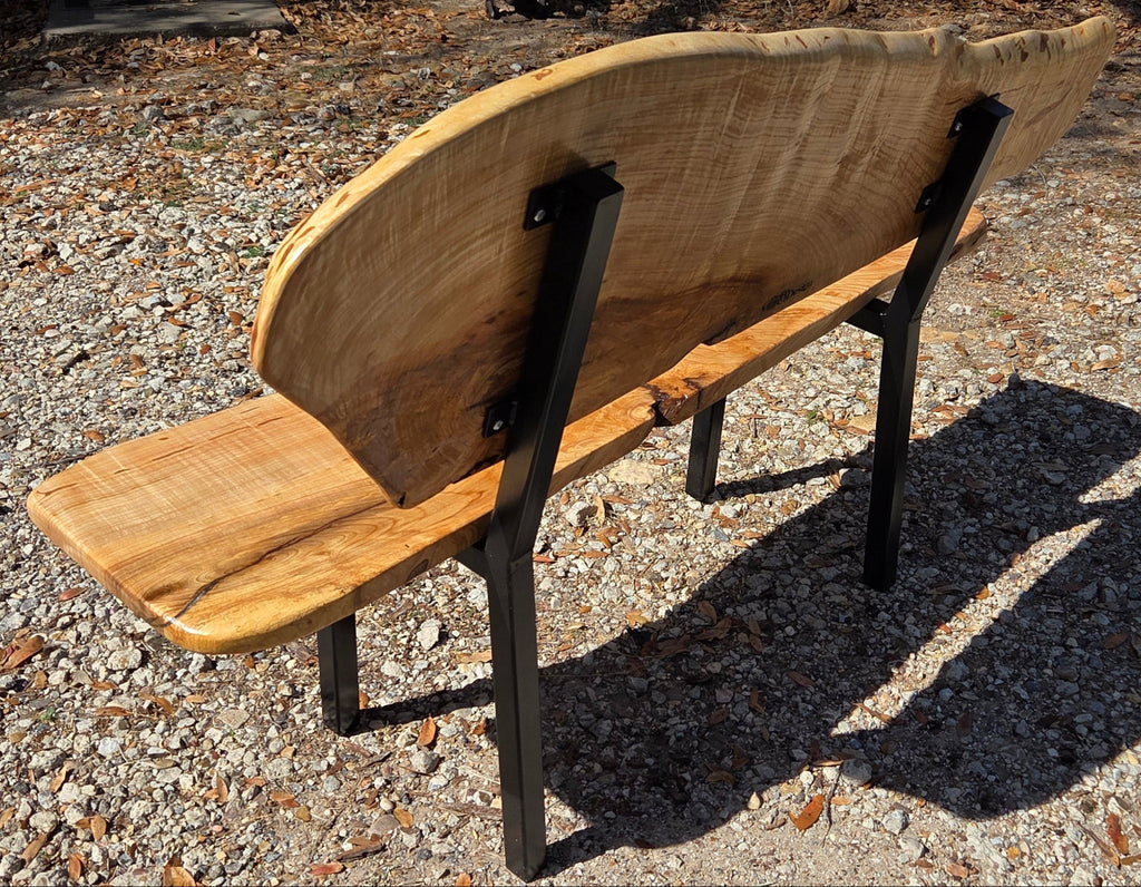 A rustic Ash live-edge wooden bench with a thick natural wood backrest and seat supported by black metal legs, photographed outdoors on gravel.