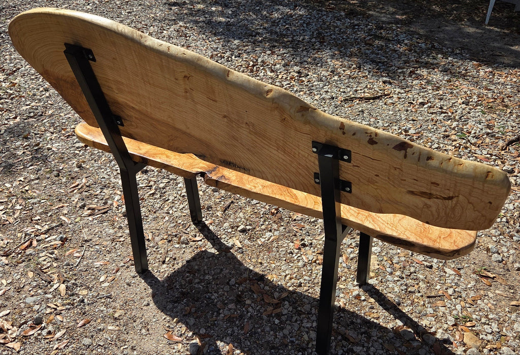 A rustic Ash live-edge wooden bench with a thick natural wood backrest and seat supported by black metal legs, photographed outdoors on gravel.
