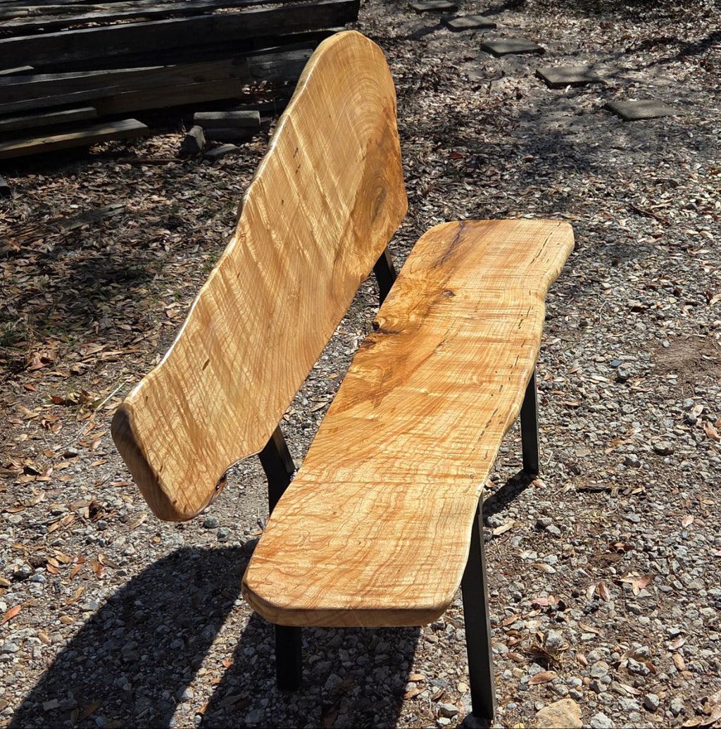 A rustic Ash live-edge wooden bench with a thick natural wood backrest and seat supported by black metal legs, photographed outdoors on gravel.