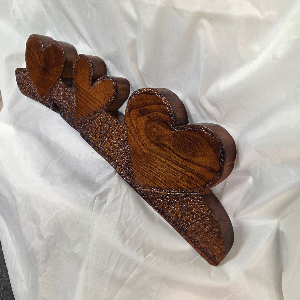 A rustic Bois D'Arc wooden plaque with three stacked heart-shaped tiers and hand carved, displayed on a wrinkled white fabric background.