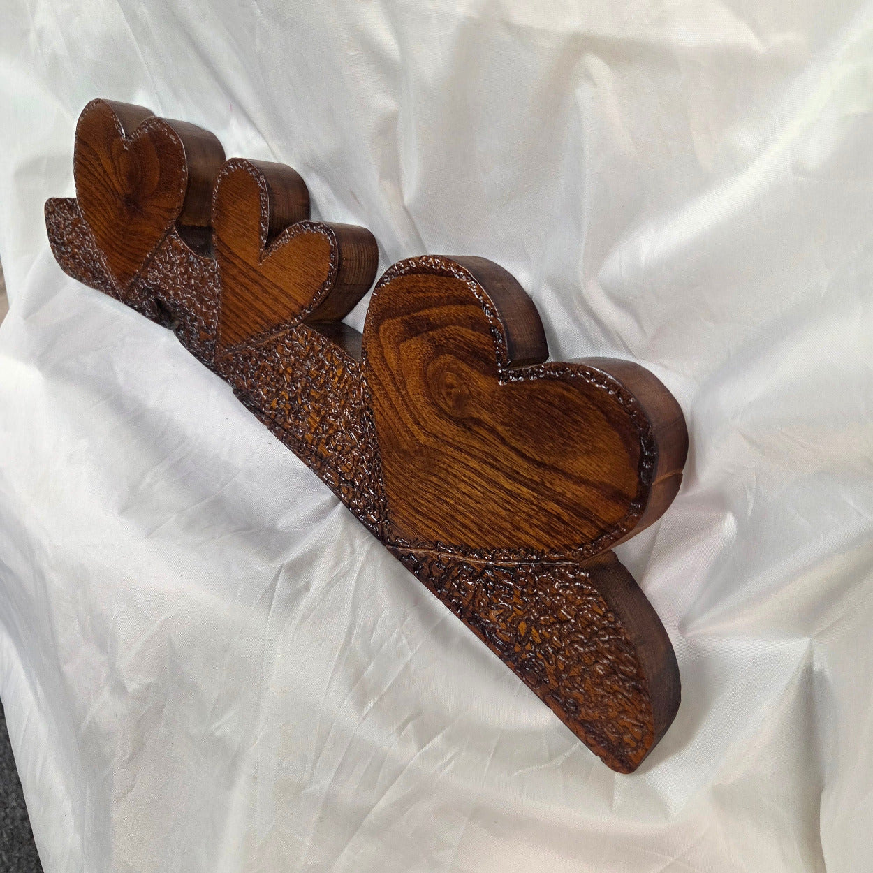 A rustic Bois D'Arc wooden plaque with three stacked heart-shaped tiers and hand carved, displayed on a wrinkled white fabric background.