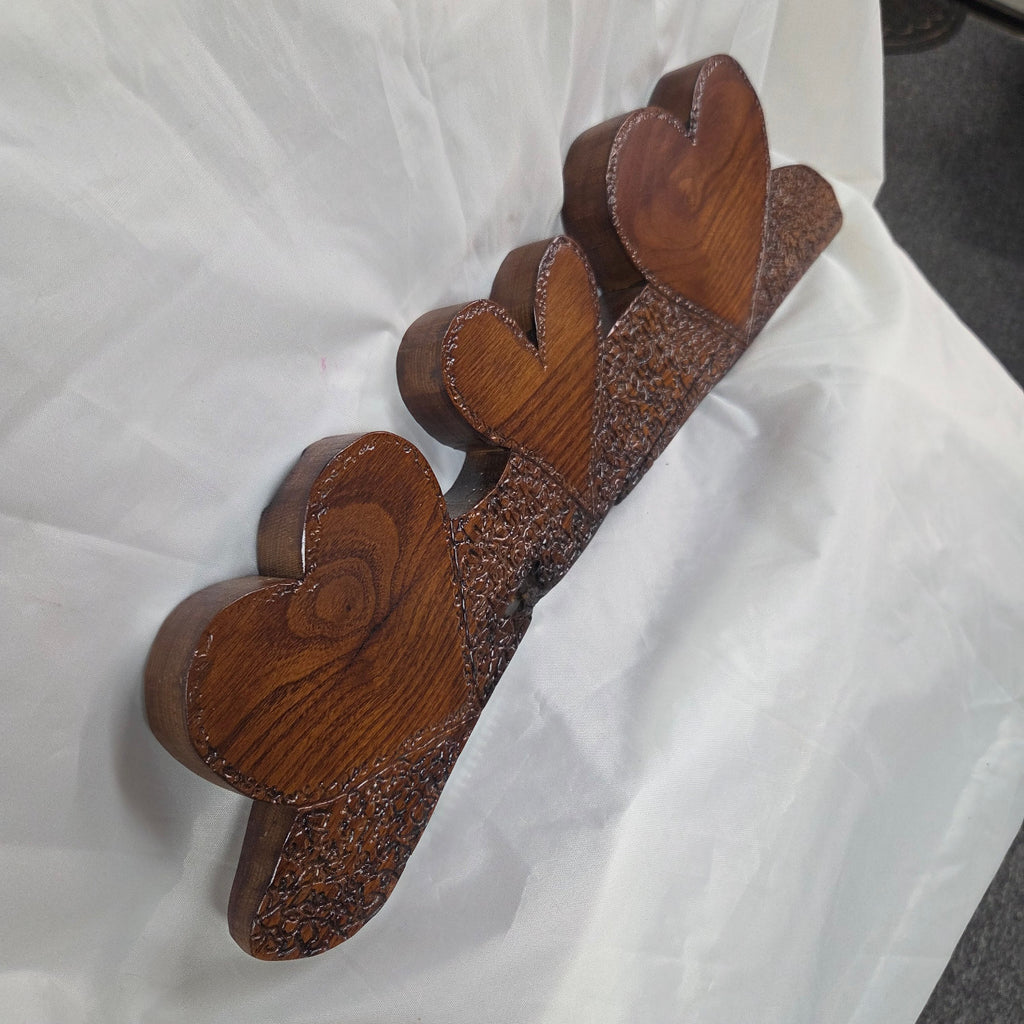 A rustic Bois D'Arc wooden plaque with three stacked heart-shaped tiers and hand carved, displayed on a wrinkled white fabric background.