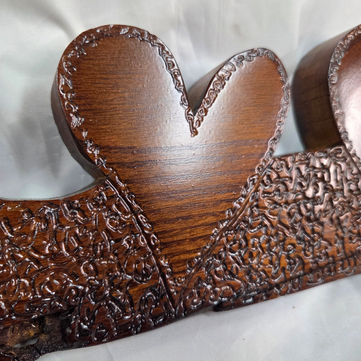 close up of A rustic Bois D'Arc wooden plaque with three stacked heart-shaped tiers and hand carved, displayed on a wrinkled white fabric background.