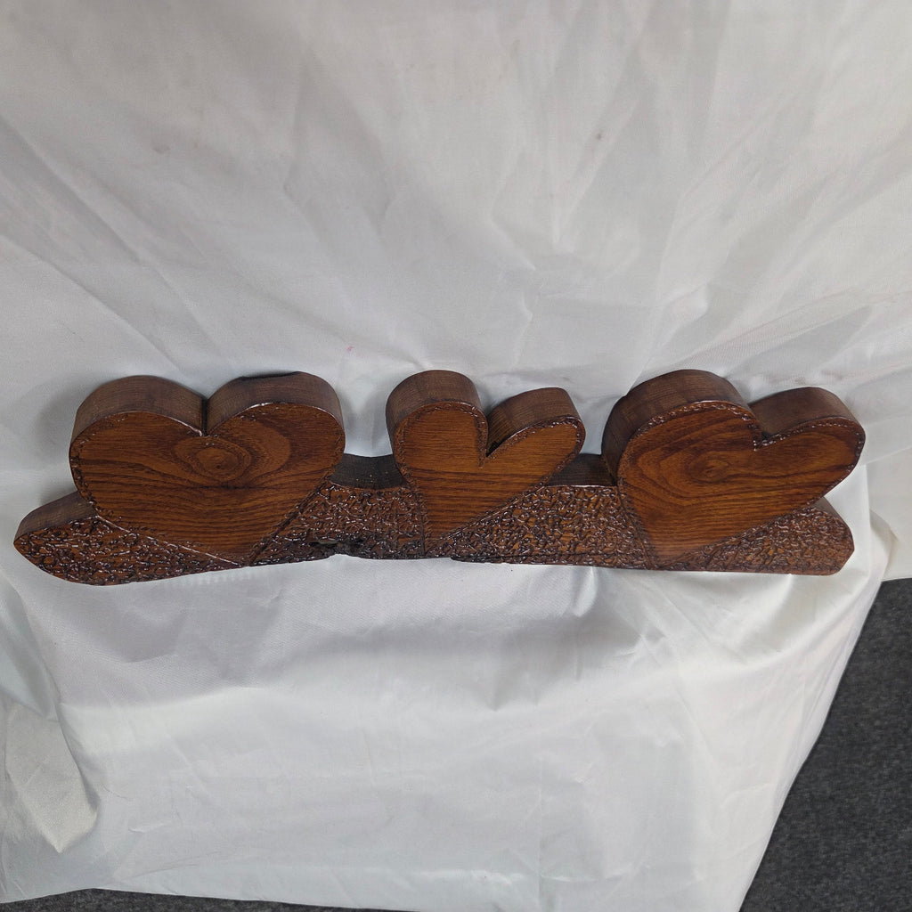 A three-heart wooden wall decor piece with a warm brown finish, displayed on a draped white fabric backdrop.