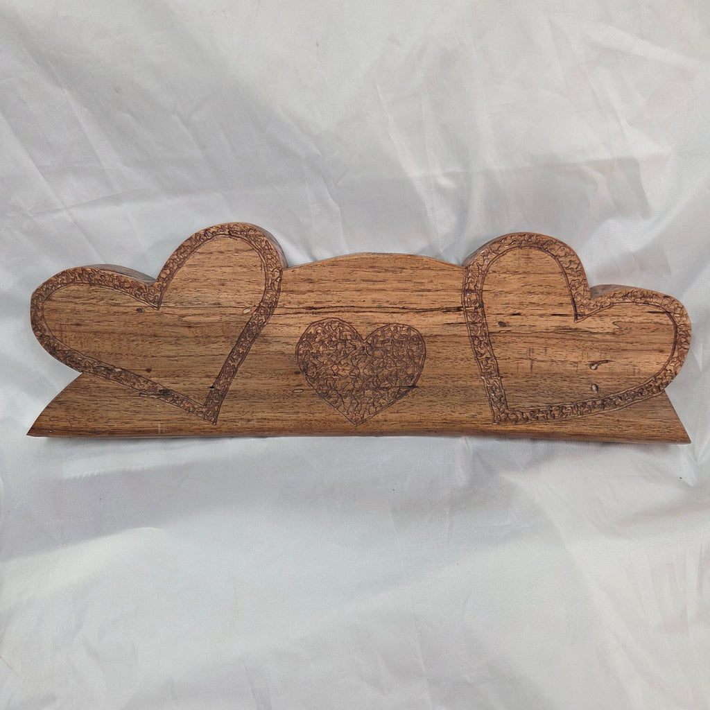 A handcrafted wooden plaque featuring three stacked heart shapes with engraved details, displayed on a wrinkled white fabric background.