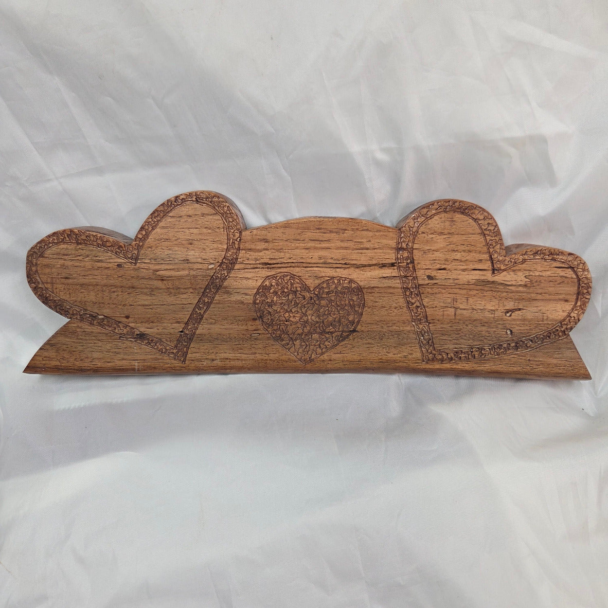 A handcrafted wooden plaque featuring three stacked heart shapes with engraved details, displayed on a wrinkled white fabric background.