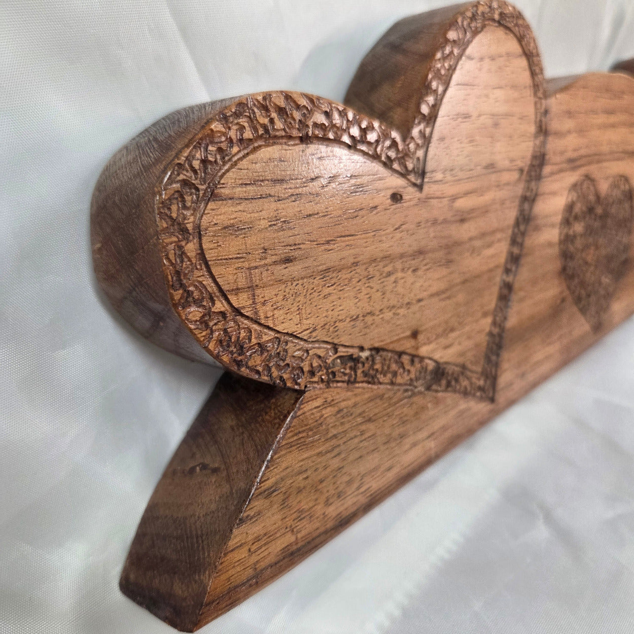 A close-up of a carved wooden heart-shaped decorative object with a smooth polished surface and engraved border details, resting on a white fabric background.
