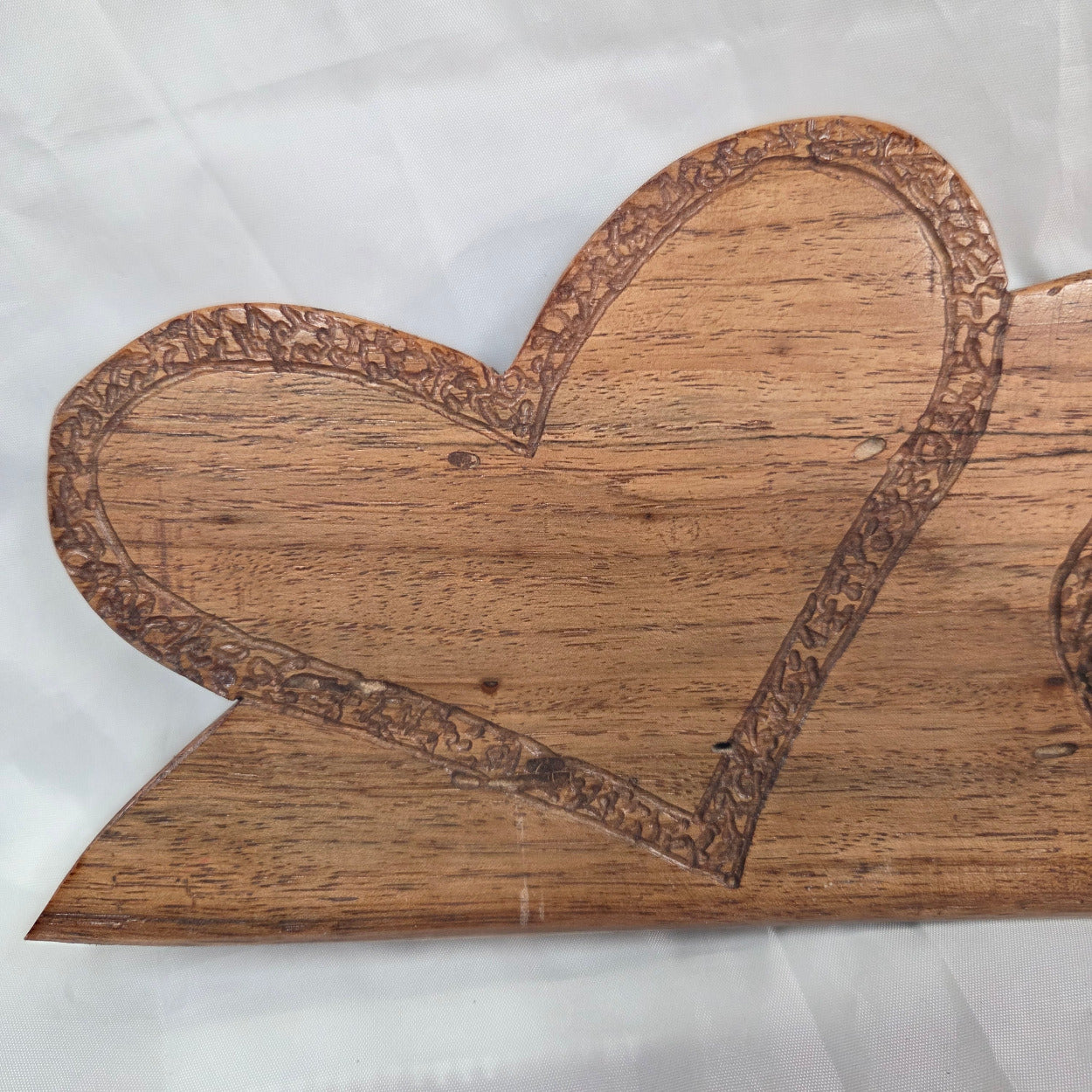 A close-up of a heart-shaped wooden plaque or wall decor with a carved floral border, showing natural wood grain and a warm brown color, laid on a white fabric background.