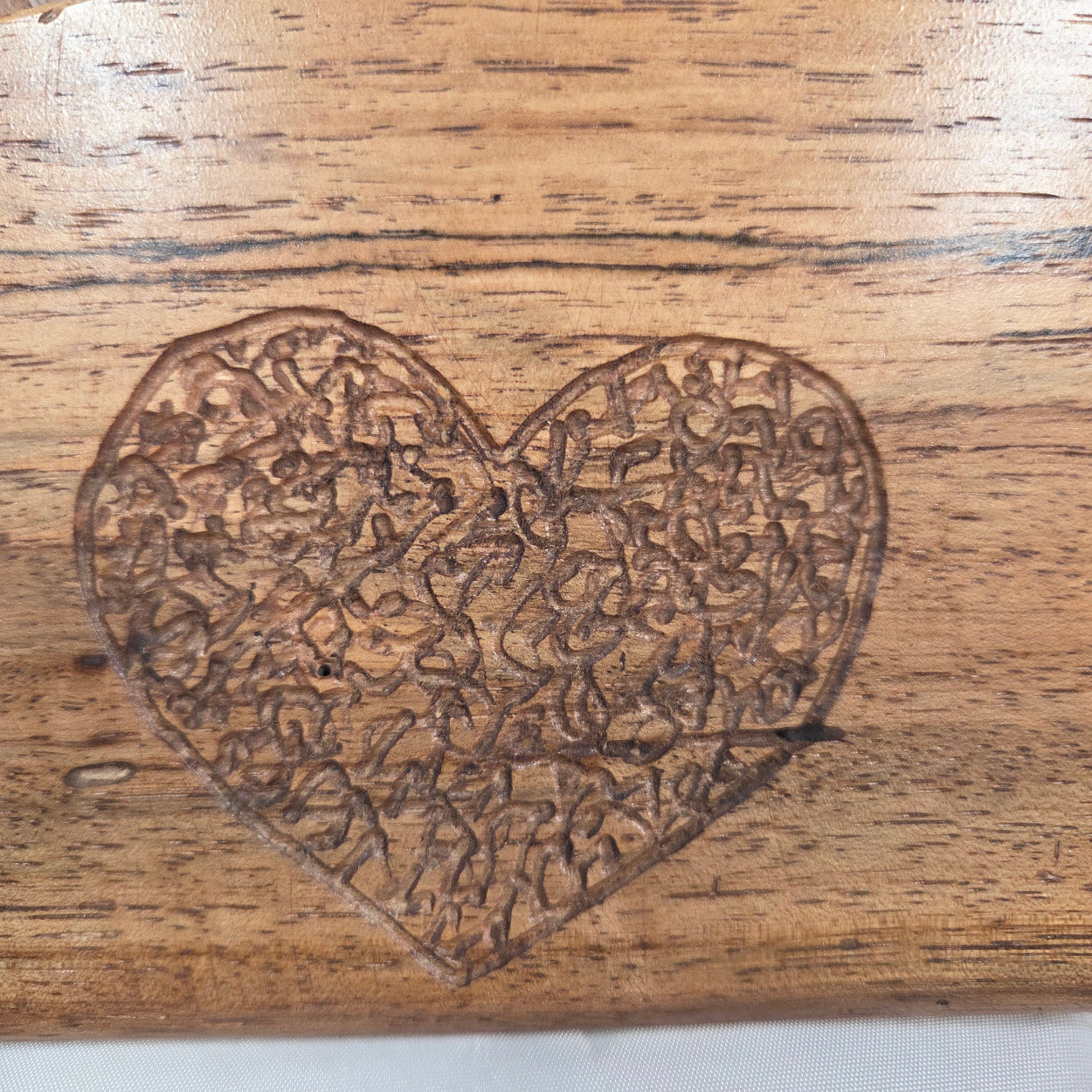 A close-up of a heart-shaped wooden engraving with an intricate floral or scroll pattern, set on a natural wood surface.