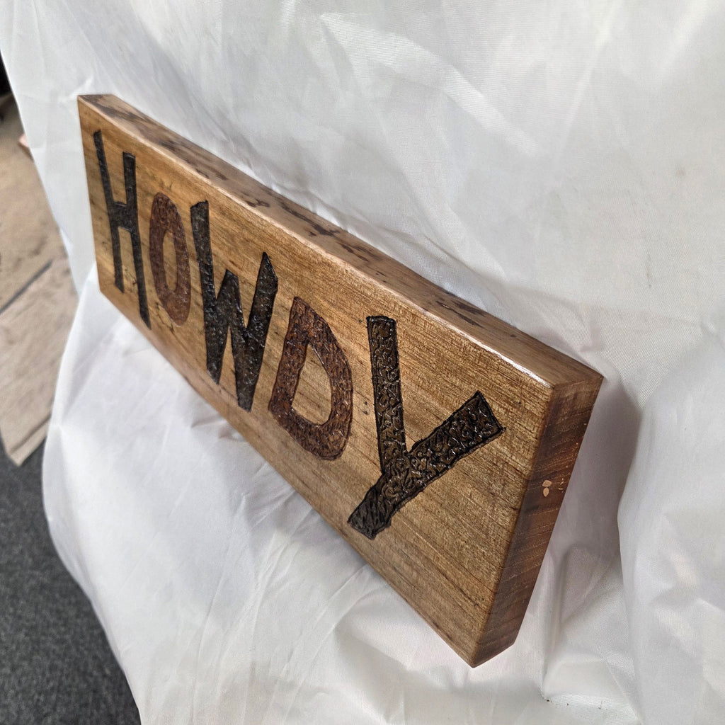 A rustic rectangular pecan sign with the word 'HOWDY' hand carved in dark brown burnt lettering,