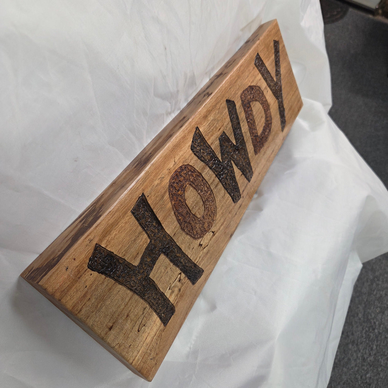A rustic rectangular pecan sign with the word 'HOWDY' hand carved in dark brown burnt lettering,