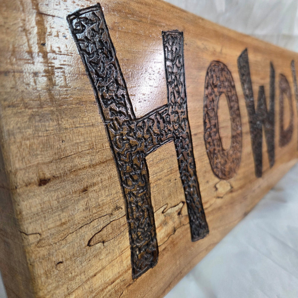A rustic rectangular pecan sign with the word 'HOWDY' hand carved in dark brown burnt lettering,