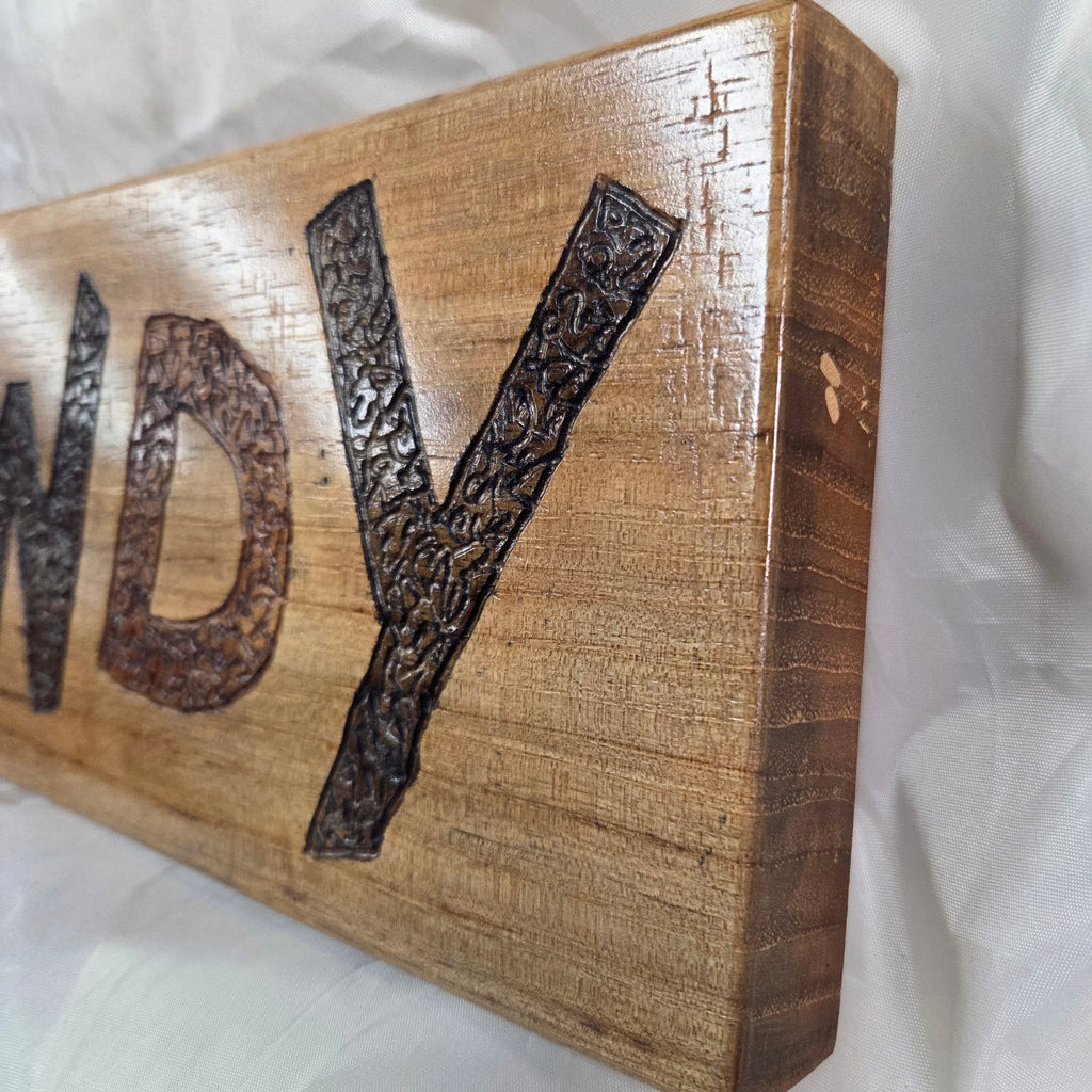 A rustic rectangular pecan sign with the word 'HOWDY' hand carved in dark brown burnt lettering,