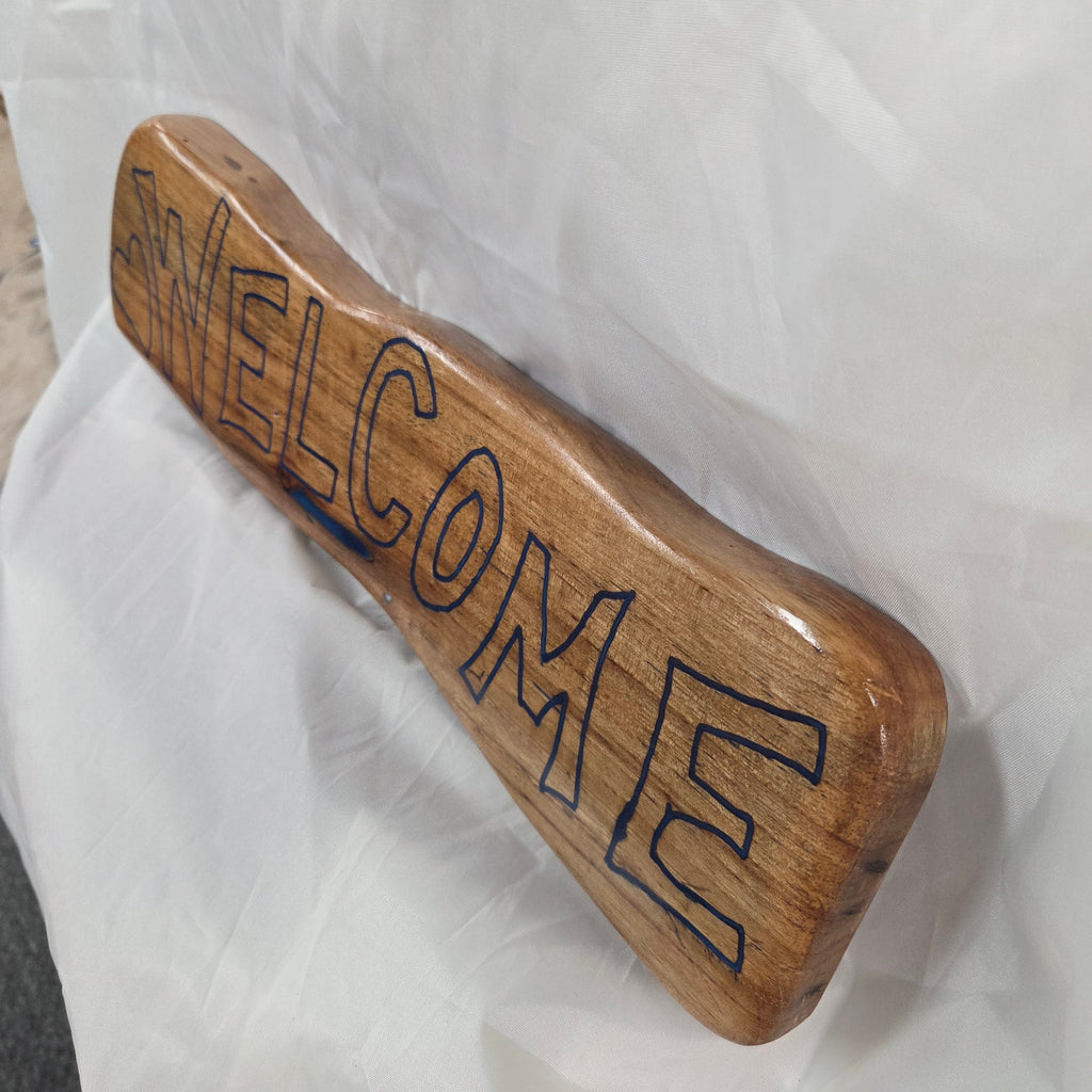 A rustic, live-edge white oak sign featuring the word 'WELCOME' in a blue epoxy outline font carved into the natural wood grain, 