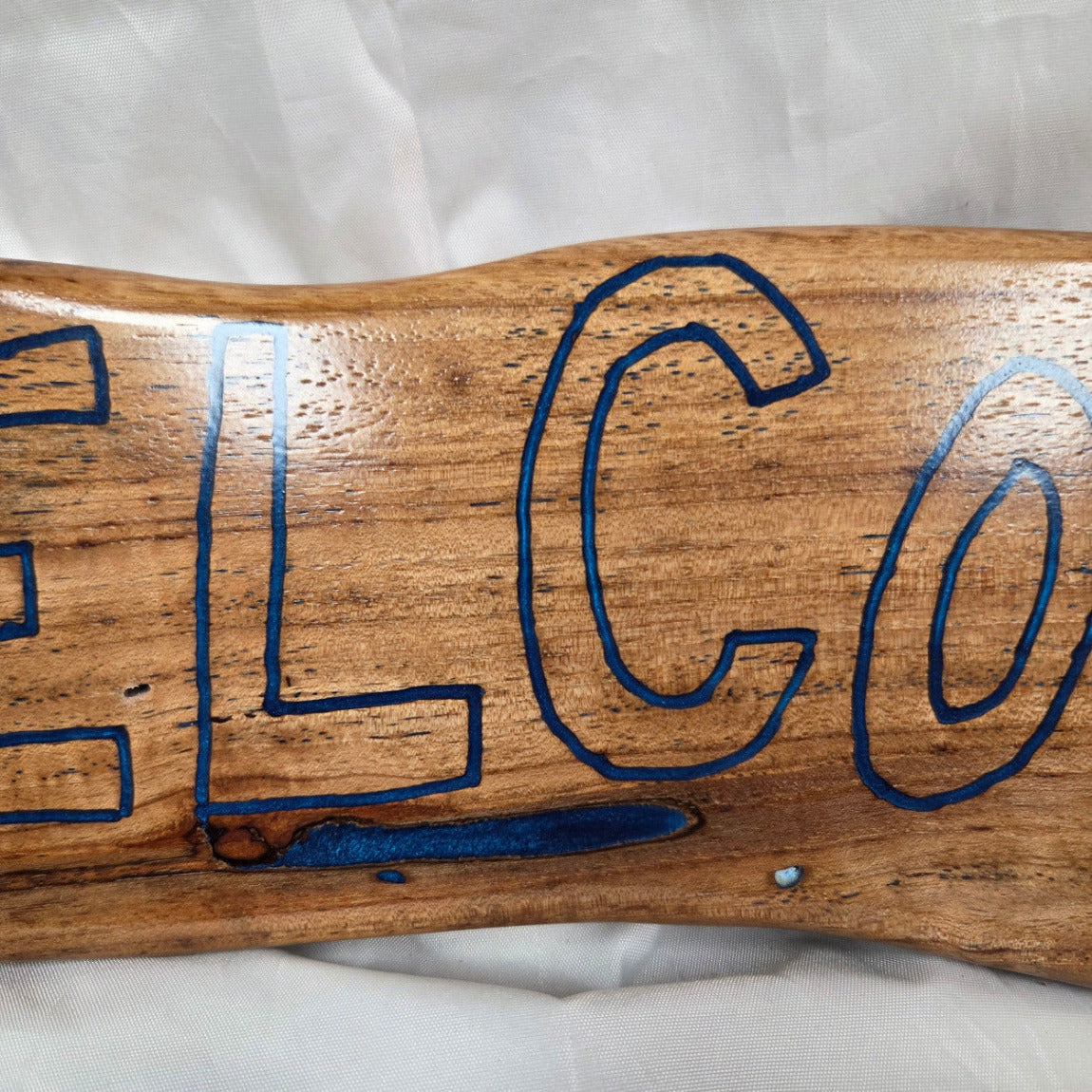 A rustic, live-edge white oak sign featuring the word 'WELCOME' in a blue epoxy outline font carved into the natural wood grain, 