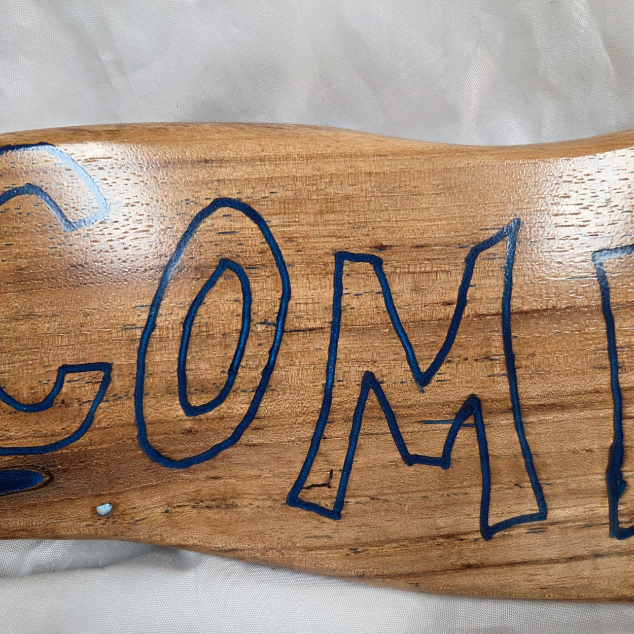 A rustic, live-edge white oak sign featuring the word 'WELCOME' in a blue epoxy outline font carved into the natural wood grain, 
