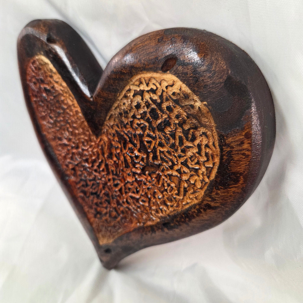 A heart-shaped wooden hanging heart with a highly textured, carved pattern on the interior, photographed on a wrinkled white fabric background.