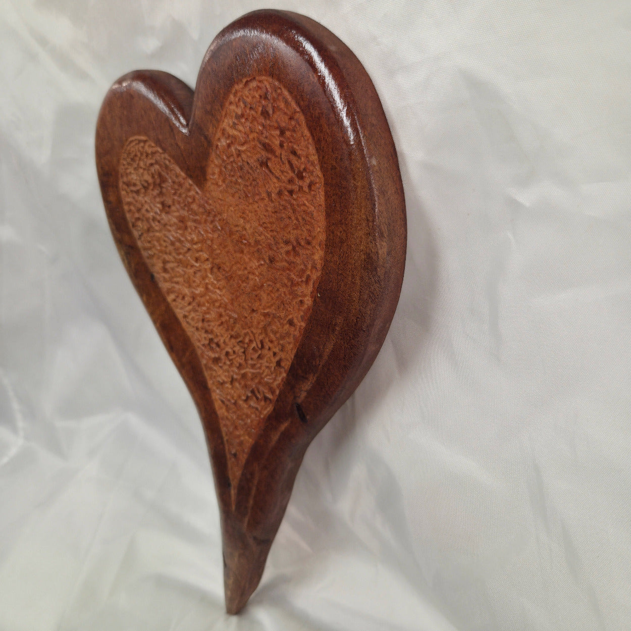 Mesquite Handing heart wall art with Hand carved center section with a smooth dark brown outer border and a lighter, textured natural wood inlay in the center, displayed against a wrinkled white fabric background.