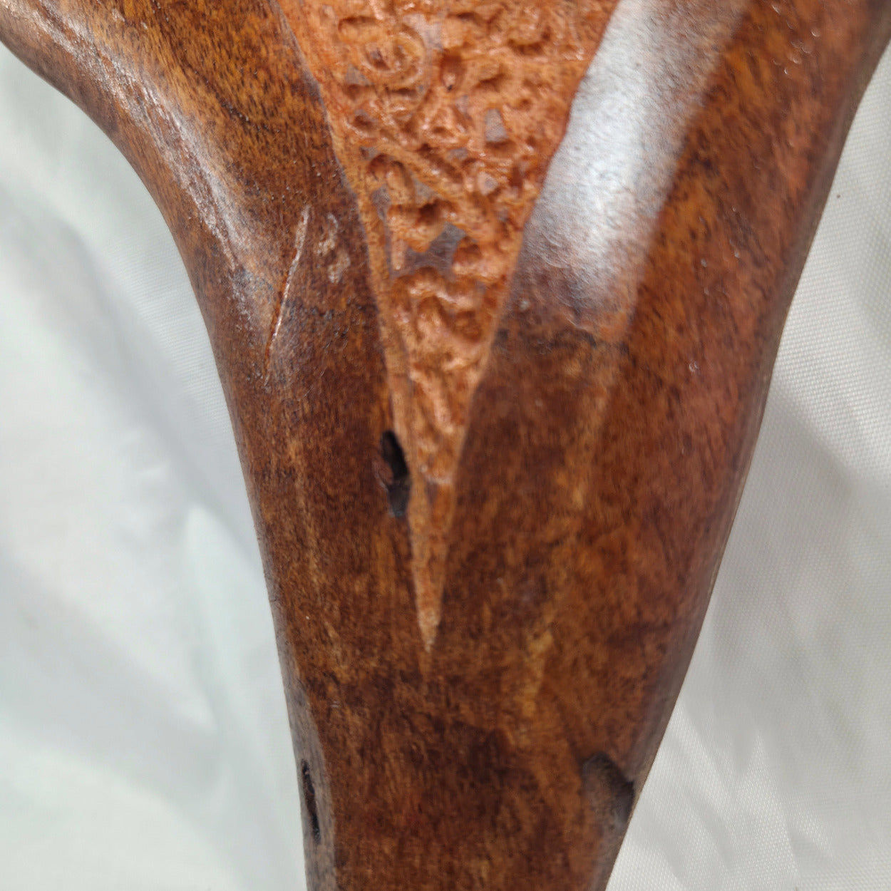 Close-up of a carved wooden object, showing rich brown wood grain, a textured carved panel, and a glossy finish against a white fabric background.
