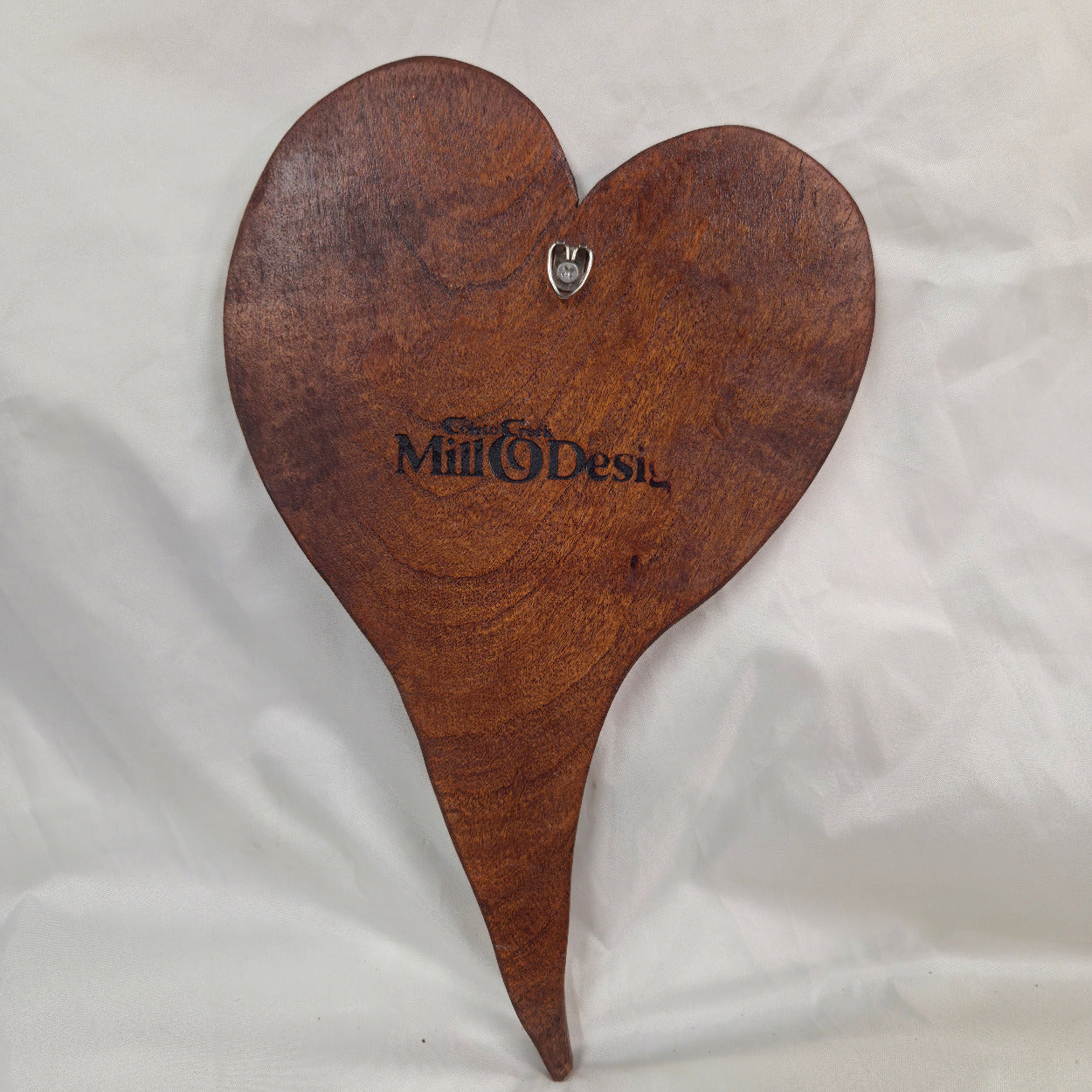 A heart-shaped wooden wall plaque with a pointed bottom tip, featuring the engraved text 'Special Mill of Desi' and a small metal hanging hook at the top, displayed on a wrinkled white fabric background.