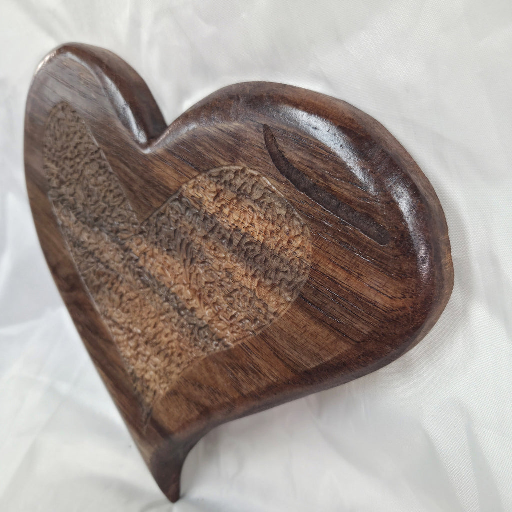 A heart-shaped wooden decorative plaque with a polished dark brown finish and a textured inlay pattern on the front, resting on a white fabric background.
