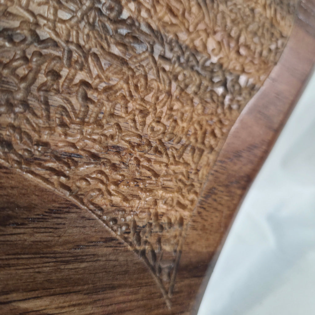 Close-up of a carved wooden surface with intricate floral or leaf-like relief patterns and a smooth wood grain border, next to a white background.