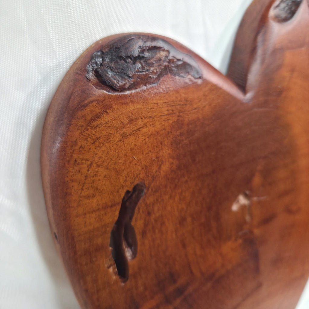 A close-up of a heart-shaped wooden plaque or wall decor with a rich reddish-brown finish, showcasing natural wood grain, knots, and a dark fissure on a white textured fabric background.