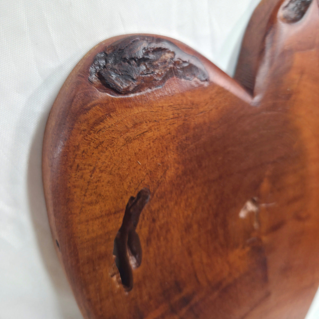 A close-up of a heart-shaped wooden plaque or wall decor with a rich reddish-brown finish, showcasing natural wood grain, knots, and a dark fissure on a white textured fabric background.