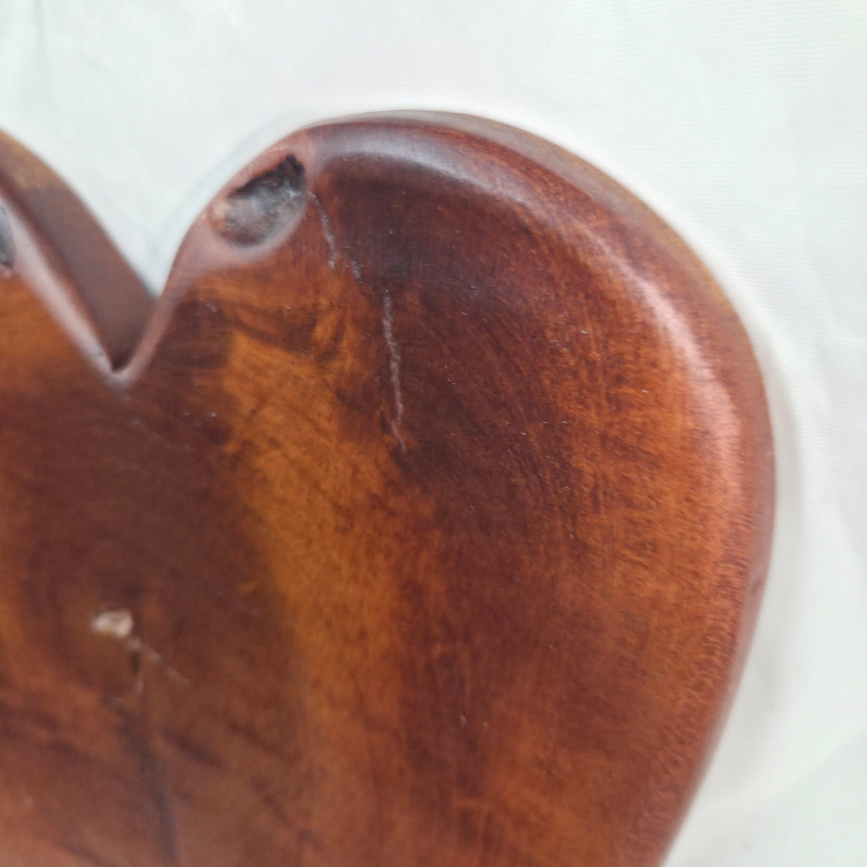 A close-up of a heart-shaped wooden object, likely a decorative plaque or carving, showing a rich reddish-brown wood grain and a small notch on the upper left edge against a plain light background.