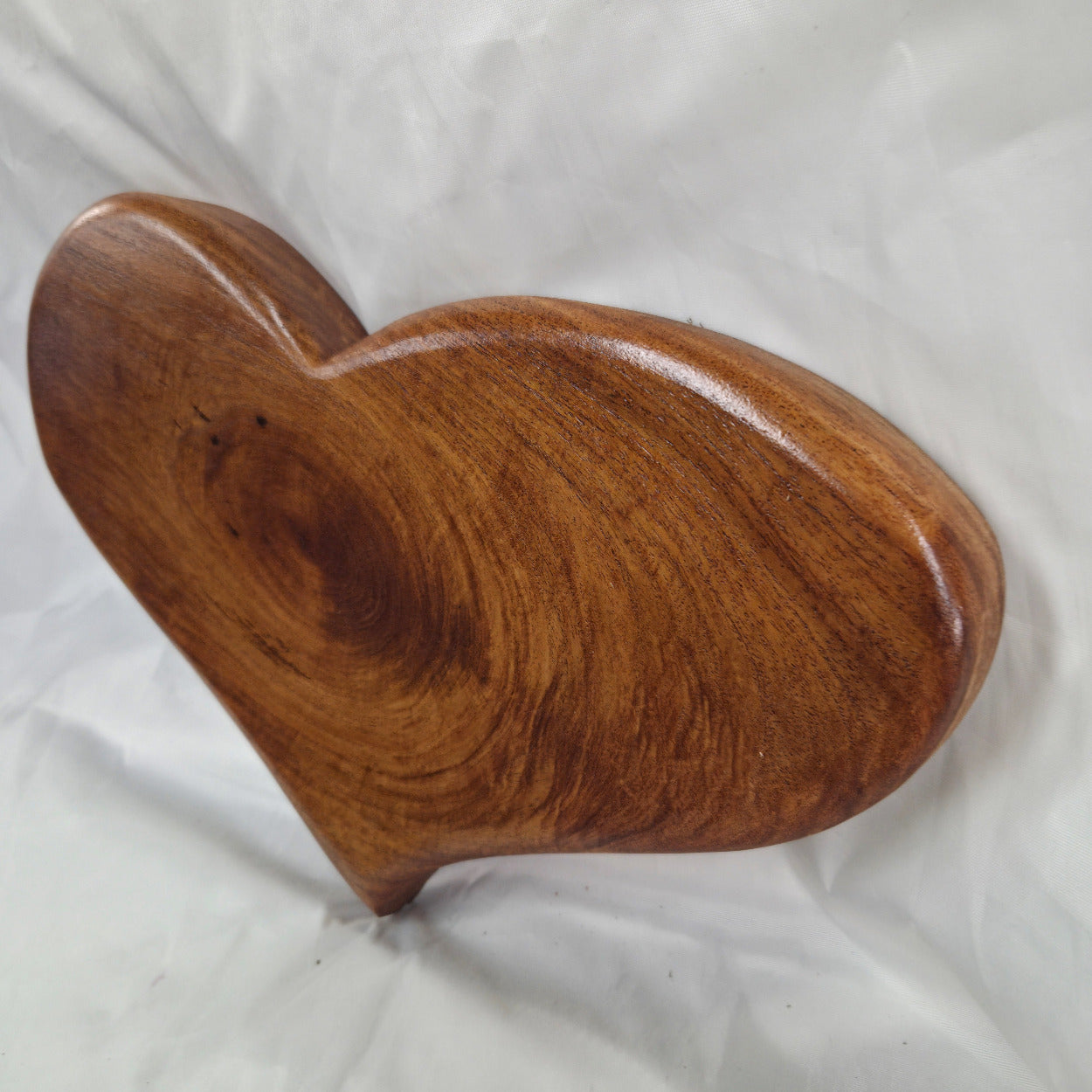 A heart-shaped wooden plaque or wall decor piece with a polished finish, showcasing rich brown wood grain patterns, set against a wrinkled white fabric background.
