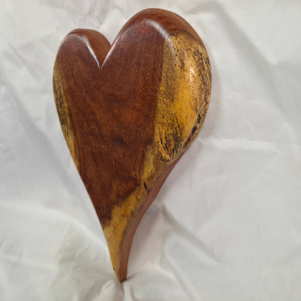 A heart-shaped wooden wall plaque with a polished finish, showcasing rich brown and golden-yellow wood grain patterns, displayed on a white fabric background.