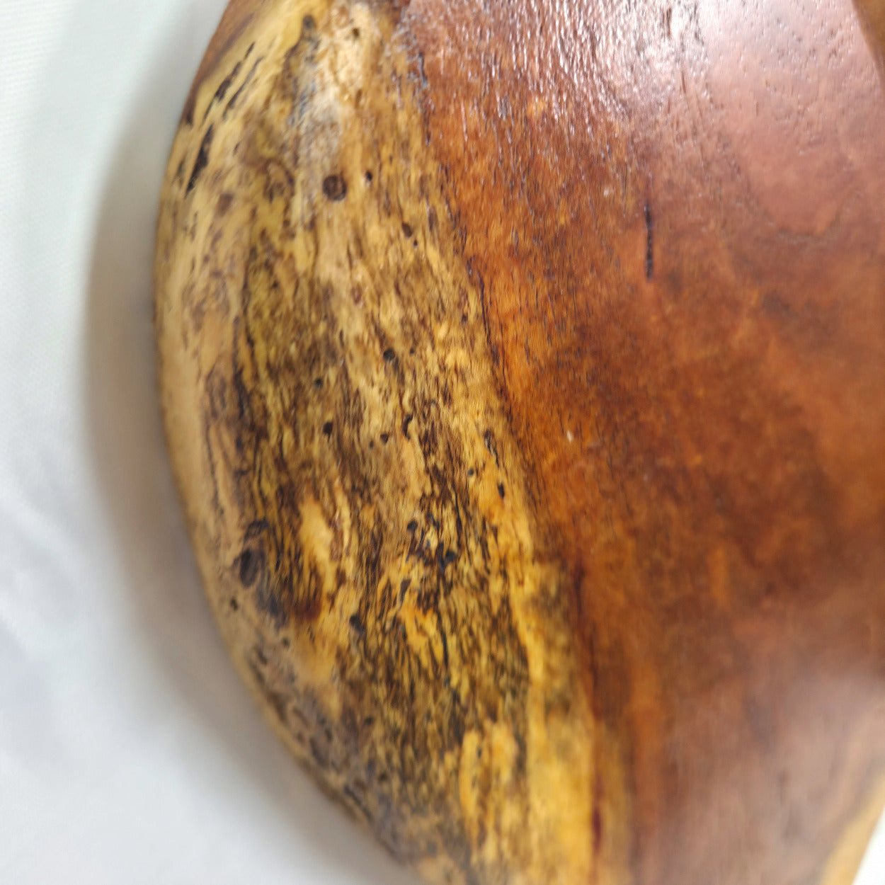 A close-up of a wooden bowl or vessel showing a natural split with one side in warm medium brown wood and the other side in lighter tan wood with dark grain patterns.