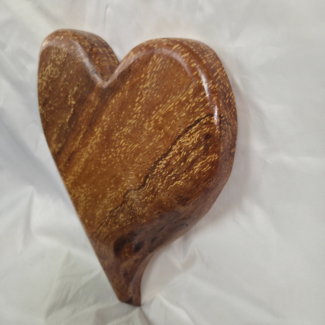 A heart-shaped White Oak Handing Heart wall art with a glossy finish and prominent natural wood grain, resting on a white fabric background.