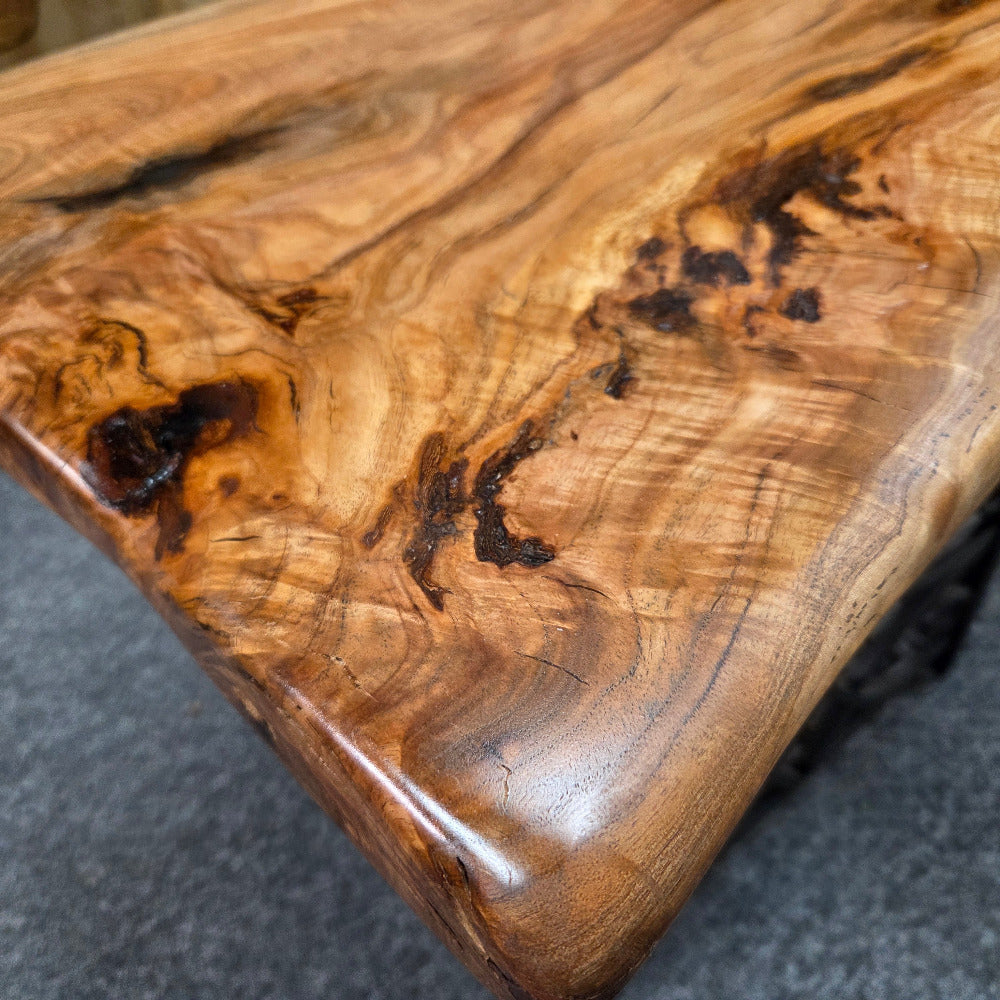 Close-up of a polished wooden tabletop with rich natural grain patterns, dark knots, and warm honey-brown tones, photographed indoors on a gray carpeted floor.