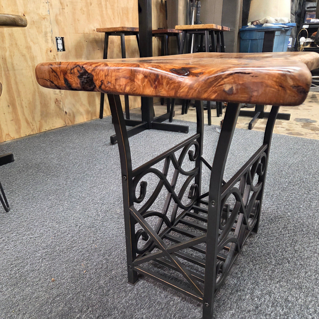 A rustic live-edge wood table with a dark metal scrollwork base, photographed in a furniture showroom on a gray carpet.