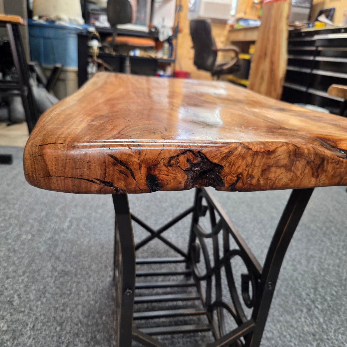 A close-up of a live-edge wooden tabletop with a glossy finish and dramatic natural wood grain, supported by a black metal frame in a workshop setting.