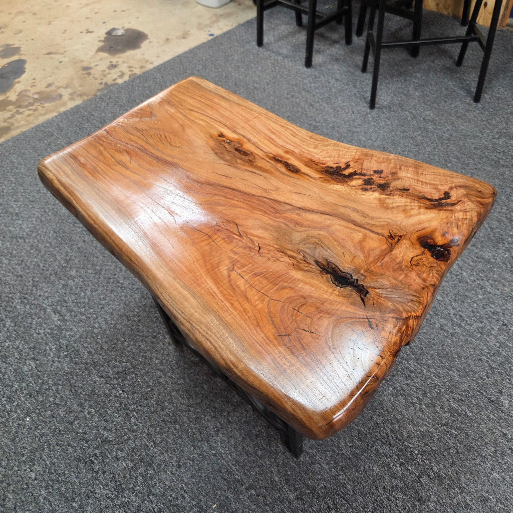 A live-edge wooden coffee table with a glossy, polished finish, displayed on a gray carpeted floor in what appears to be a showroom or warehouse.