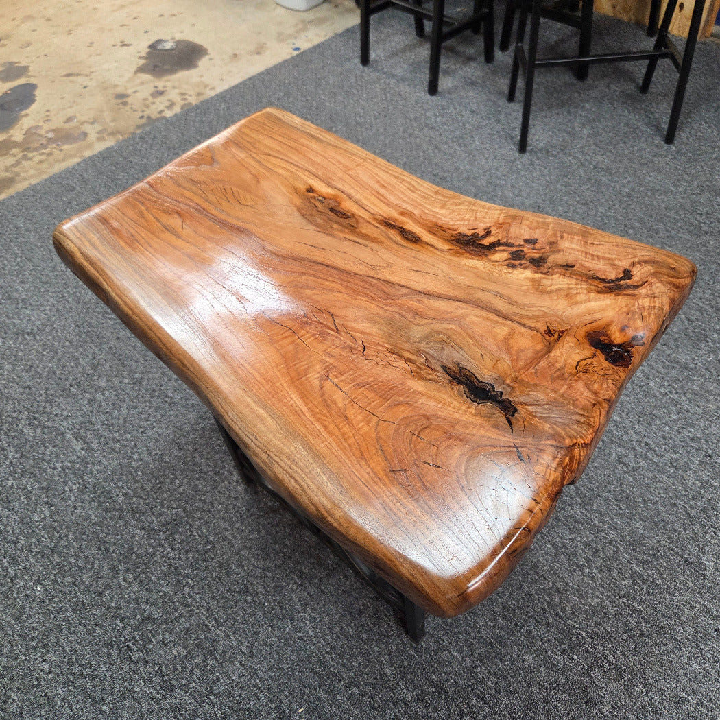 A live-edge wooden coffee table with a glossy, polished finish, displayed on a gray carpeted floor in what appears to be a showroom or warehouse.