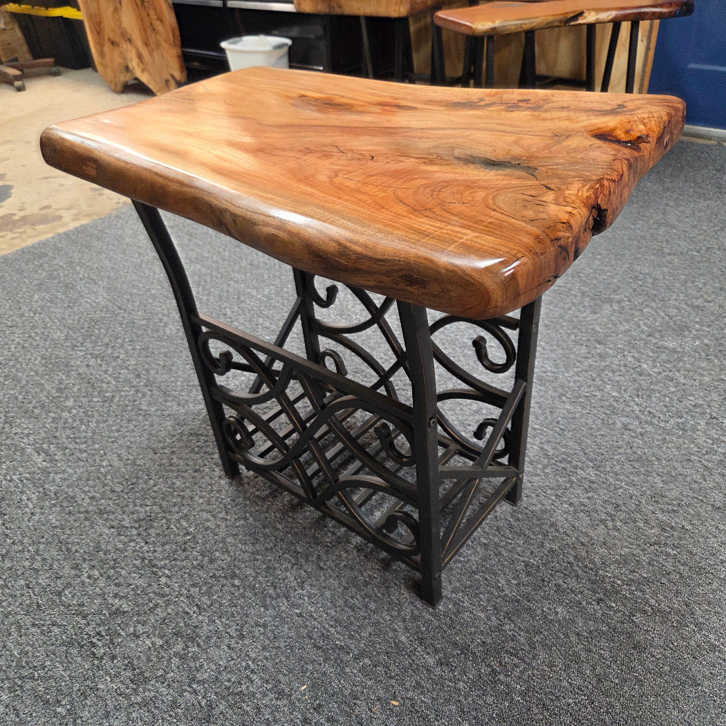 A rustic live-edge wooden table with a glossy brown top and a black wrought-iron scrollwork base, photographed on a gray carpeted floor in what appears to be a furniture showroom.