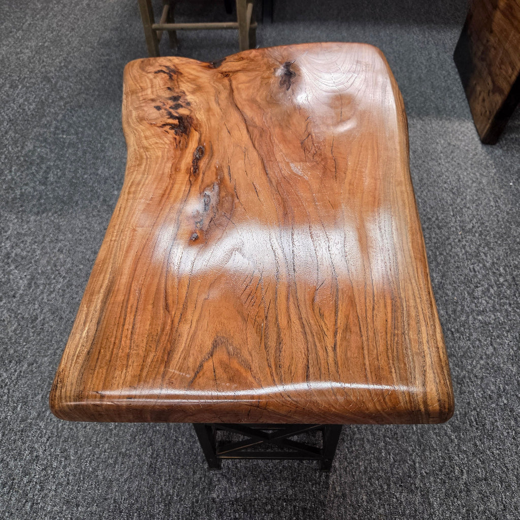 A live-edge wooden table with a glossy finish, showcasing rich natural wood grain and knots, supported by a black metal frame on a gray carpeted floor.