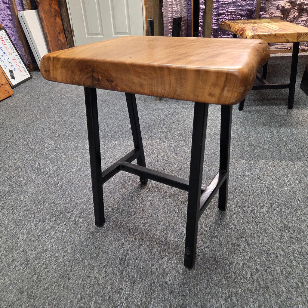 A rustic Live Oak end table with a thick, live-edge natural wood seat and a black metal frame, set on a gray carpeted floor.