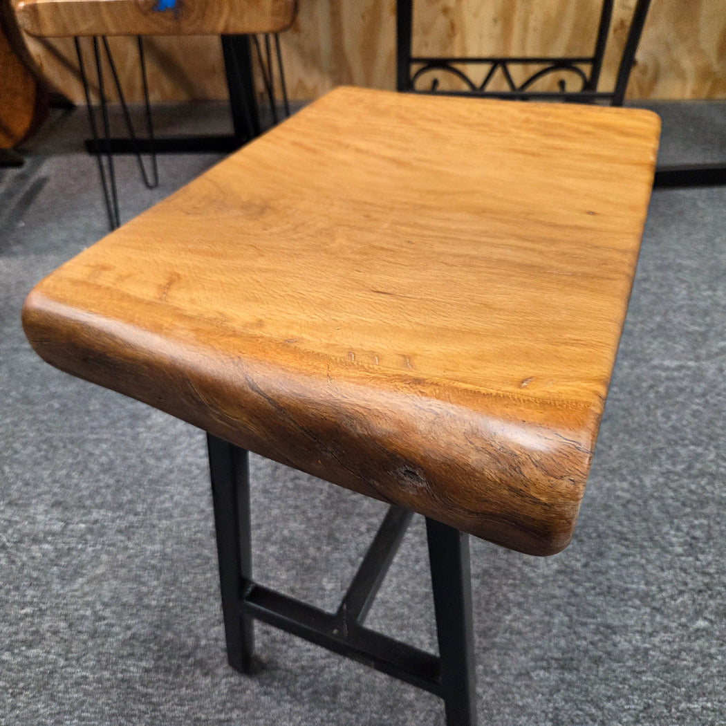 A rustic Live Oak end table with a thick, live-edge natural wood seat and a black metal frame, set on a gray carpeted floor.