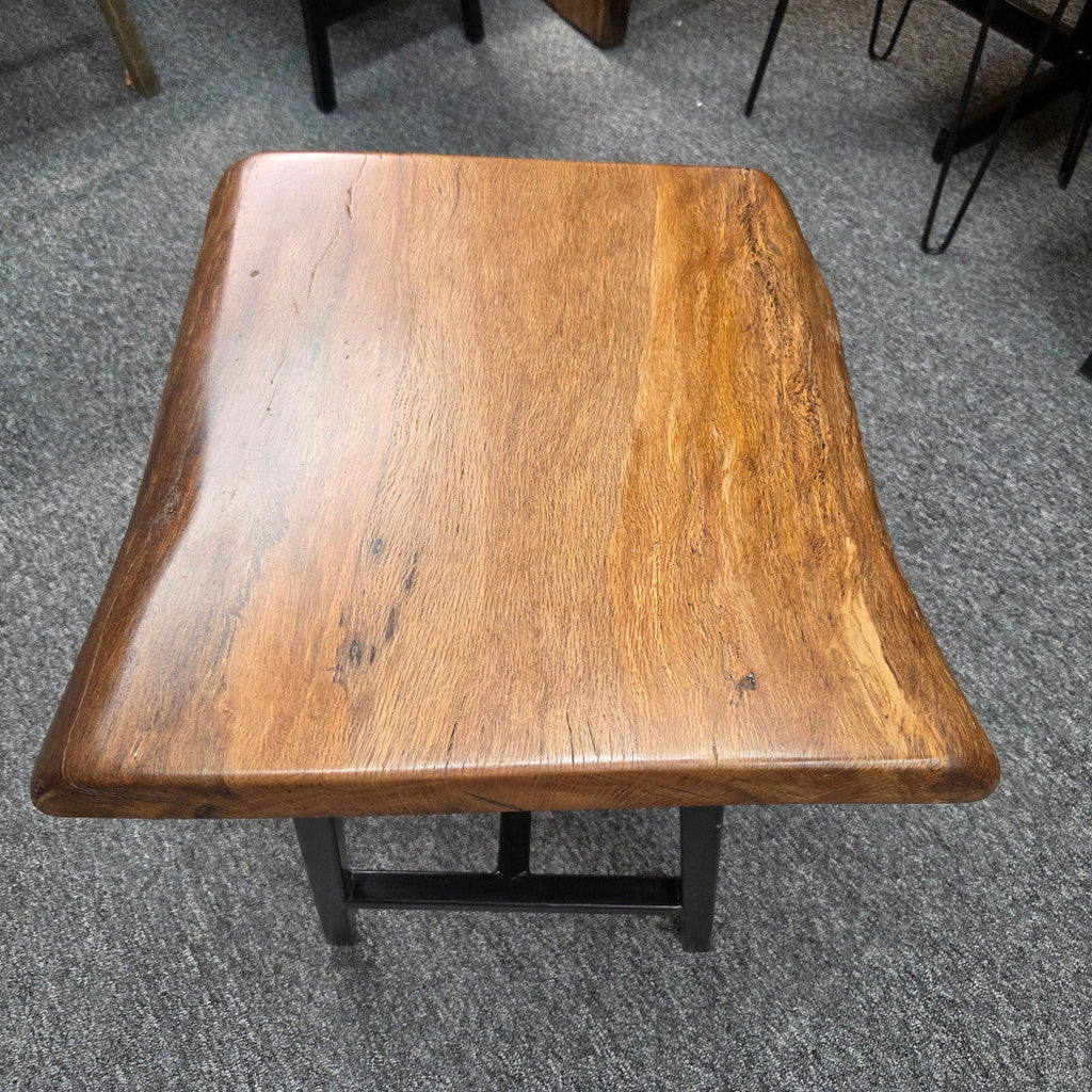 A square Live Oak end table with a live-edge natural top and black metal legs, furniture showroom setting on a gray carpet.