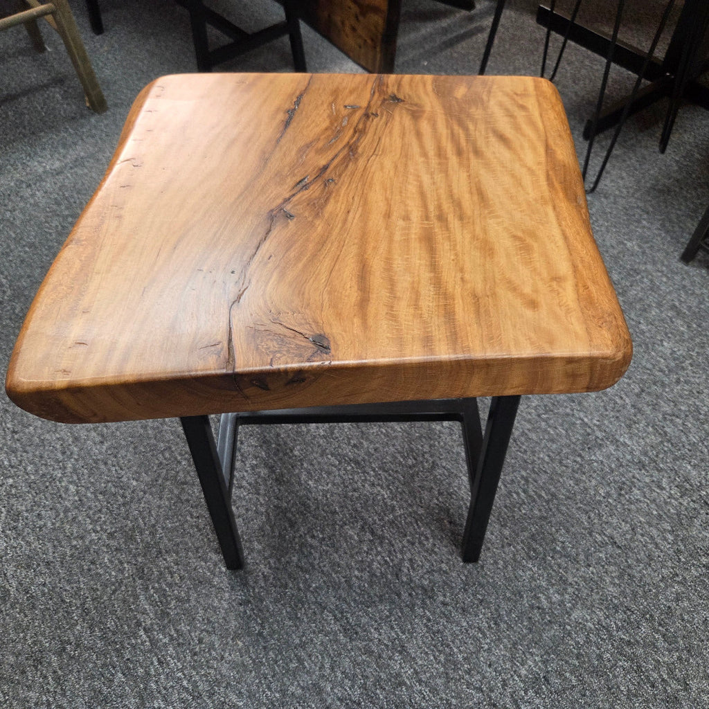 A rustic live-edge Live Oak end table with a square top and black metal legs, photographed on a gray carpeted floor.