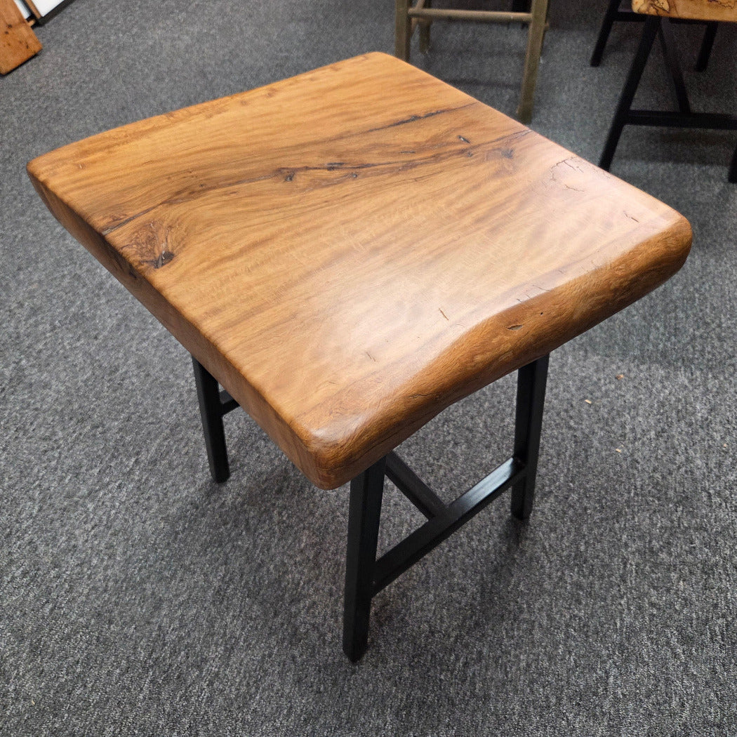 A rustic live-edge Live Oak end table with a square top and black metal legs, photographed on a gray carpeted floor.
