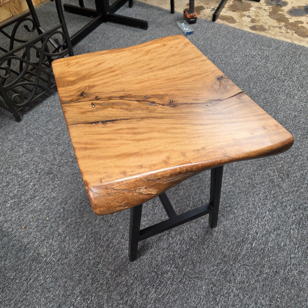 A rustic live-edge Live Oak end table with a square top and black metal legs, photographed on a gray carpeted floor.