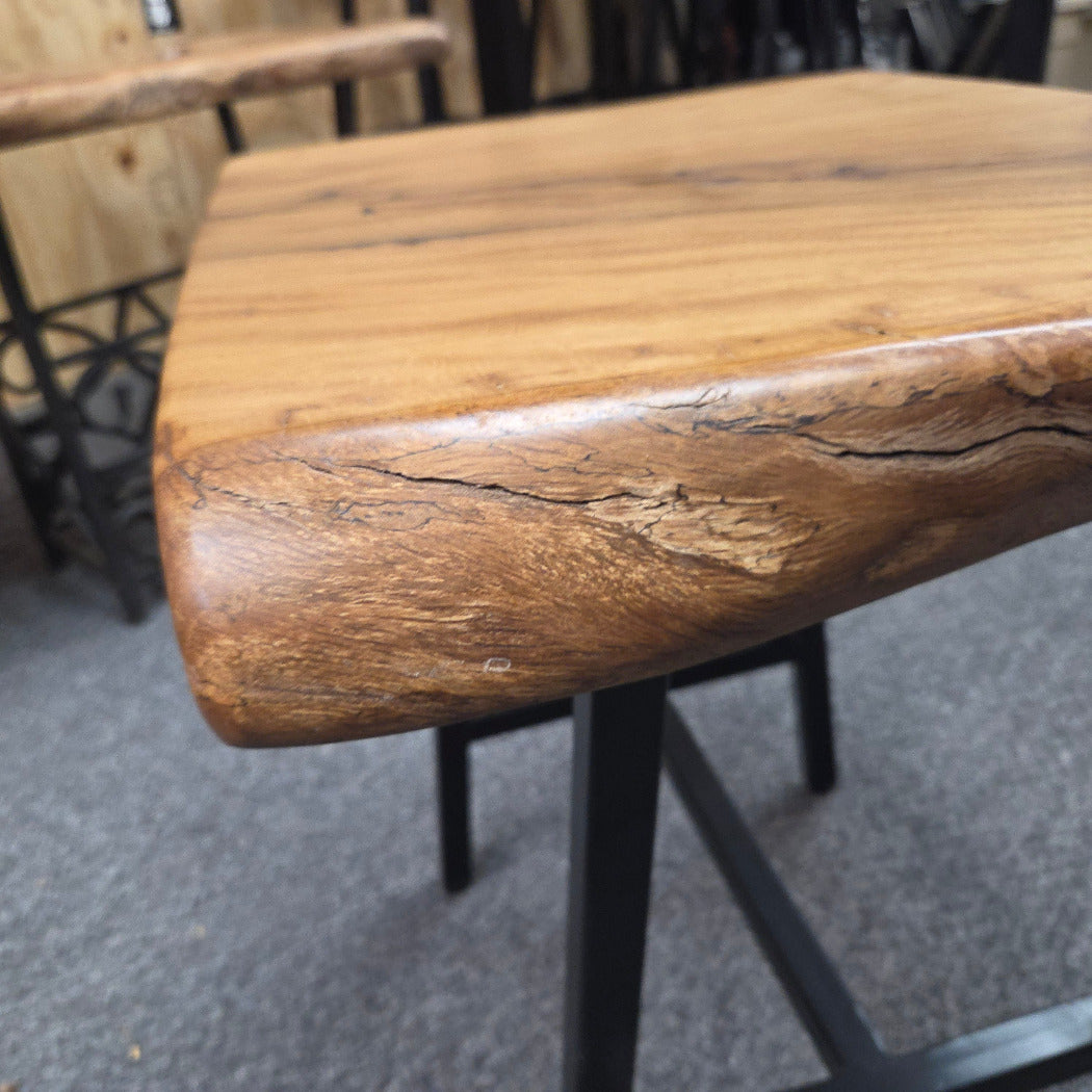 A rustic live-edge Live Oak end table with a square top and black metal legs, photographed on a gray carpeted floor.