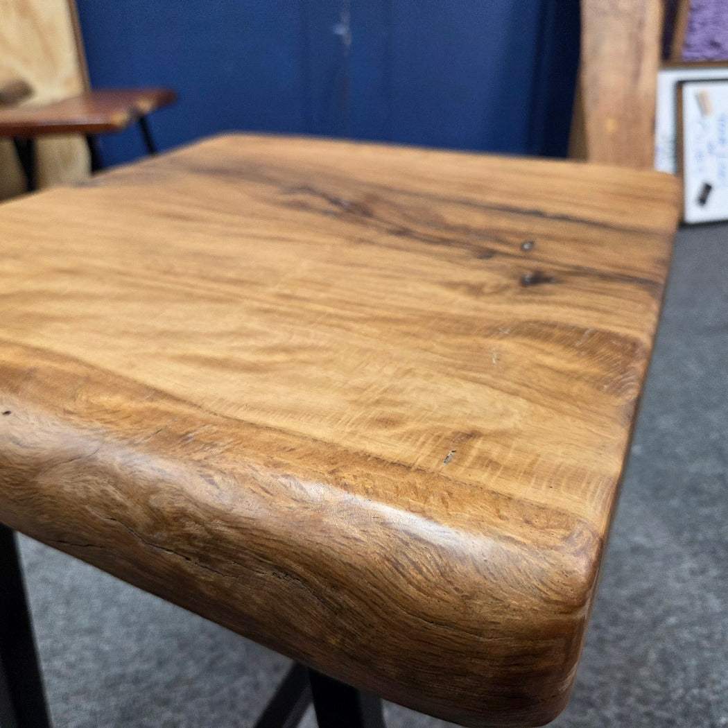 close up A rustic live-edge Live Oak end table with a square top and black metal legs, photographed on a gray carpeted floor.